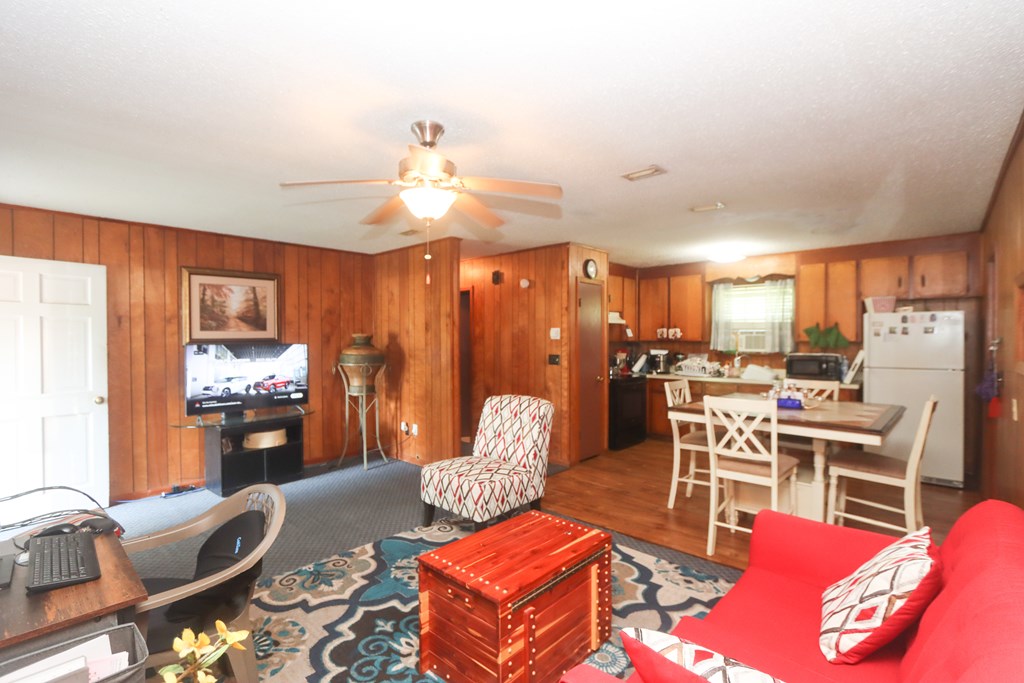 Living room with wood-paneled walls, a ceiling fan, and adjoining dining area and kitchen.