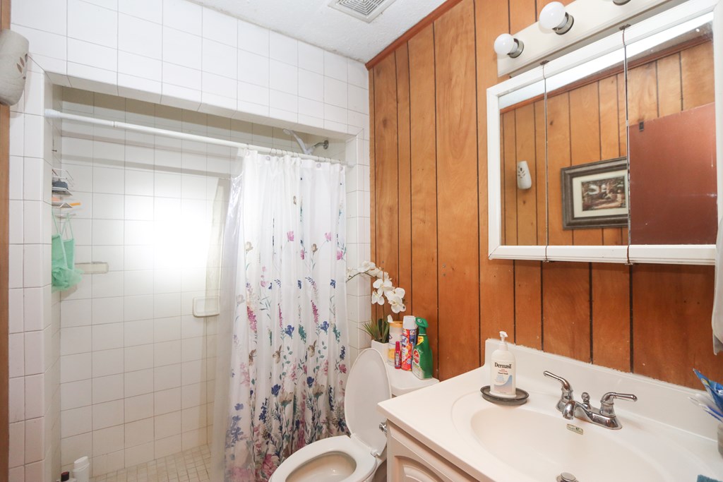 Bathroom with a tiled shower, floral curtain, wood-paneled wall, and white vanity.