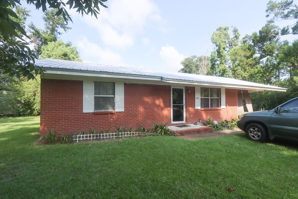 Red brick single-story house with a metal roof, white-trimmed windows, and a broad green lawn.