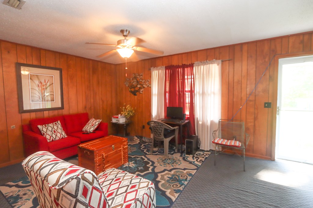 Living room with wood-paneled walls, red sofa, patterned chairs, and a desk by curtained windows.