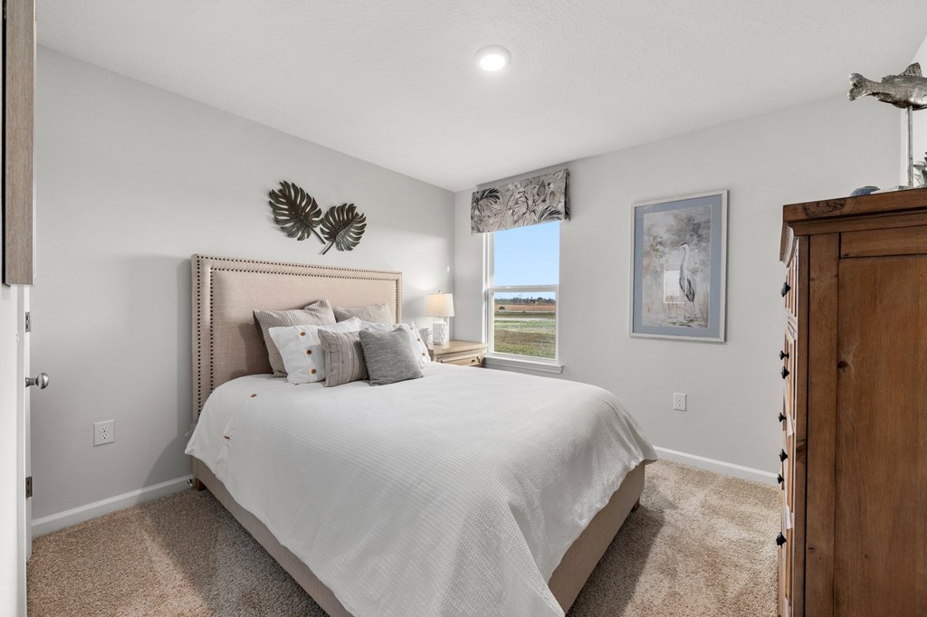 Bedroom with a white-dressed bed, beige upholstered headboard, wooden dresser, and coastal view through the window.
