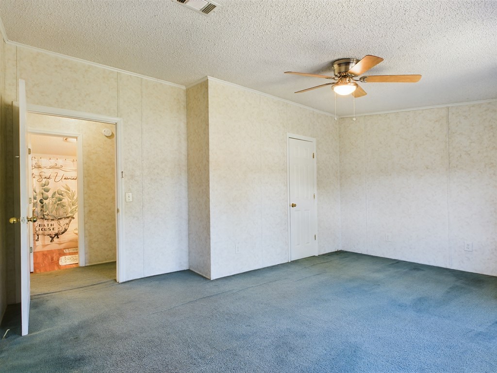 Empty bedroom with blue-green carpet, pale patterned walls, and a ceiling fan with a light.