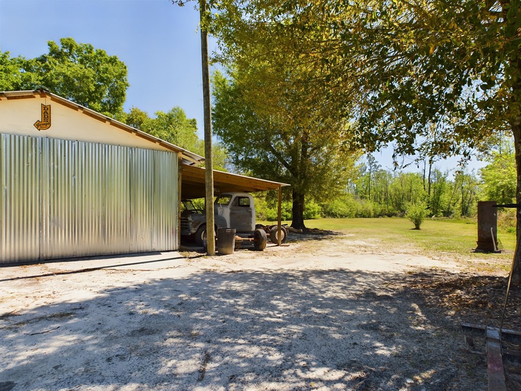 Metal-sided outbuilding with a covered parking area and old pickup truck in a sunny, wooded yard.