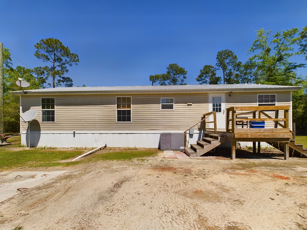 Beige manufactured home with a raised wooden deck, gravel driveway, and tall trees.