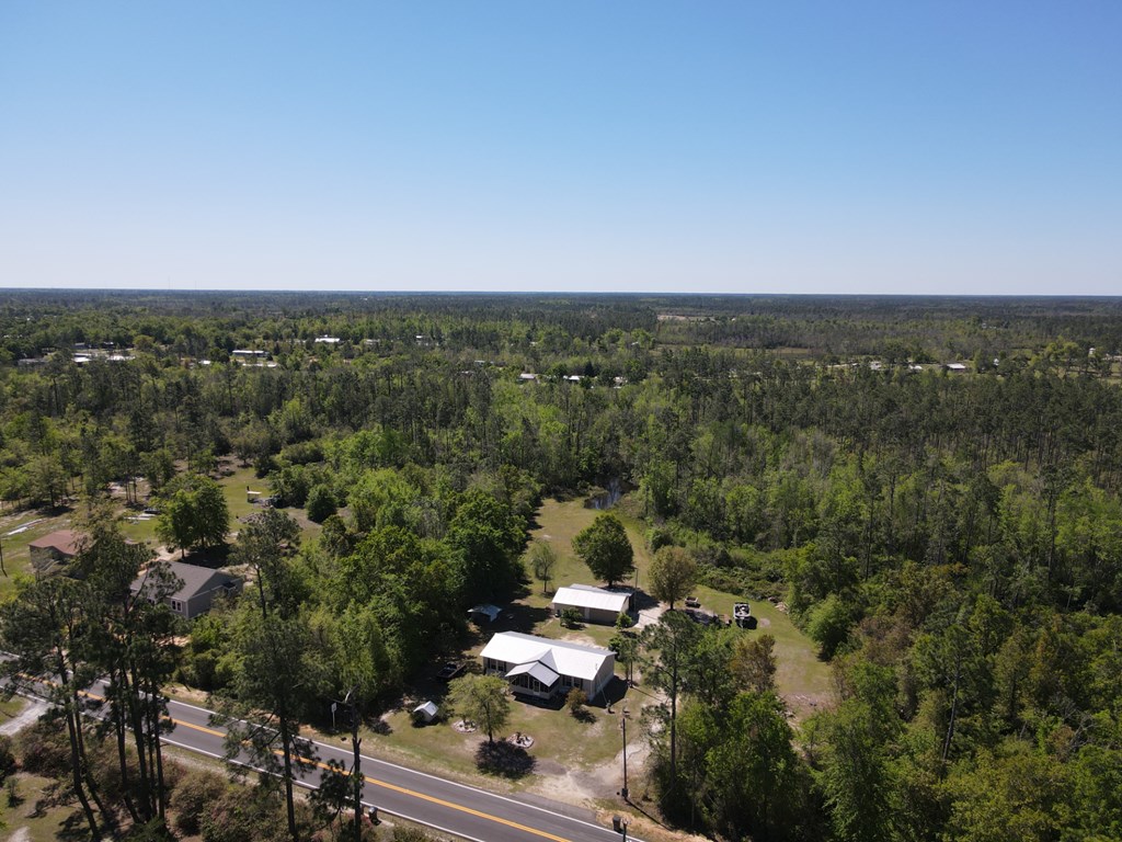 Aerial view of a rural home with white roofs, outbuildings, open yard, and surrounding trees.