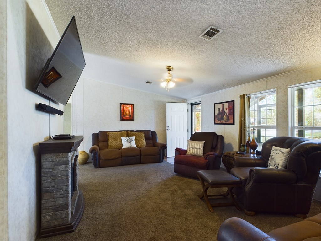 Furnished living room with brown leather seating, a stone fireplace, wall-mounted television, and bright windows.
