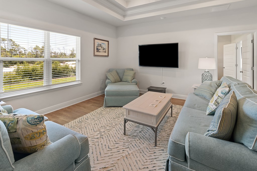 Living room with pale blue seating, a wall-mounted television, and large windows with white blinds.