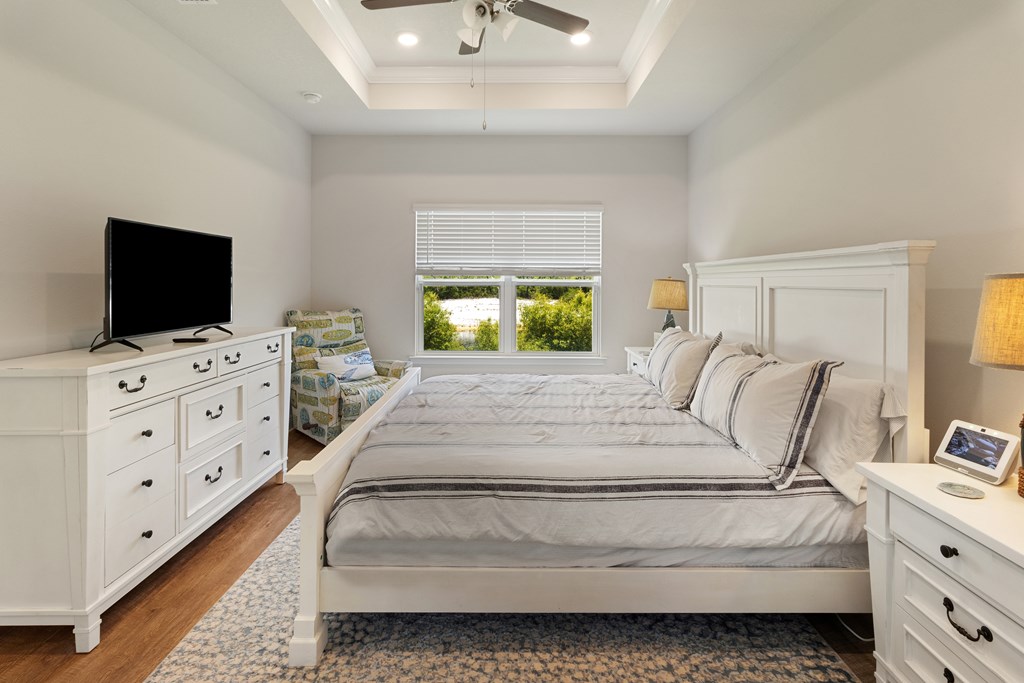 Bedroom with a large white bed, tray ceiling, and window overlooking greenery.