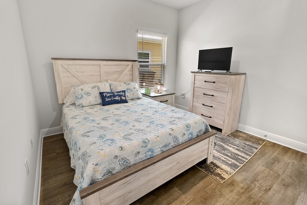 Furnished bedroom with a light wood bed, patterned coastal bedding, and a dresser topped with a television.