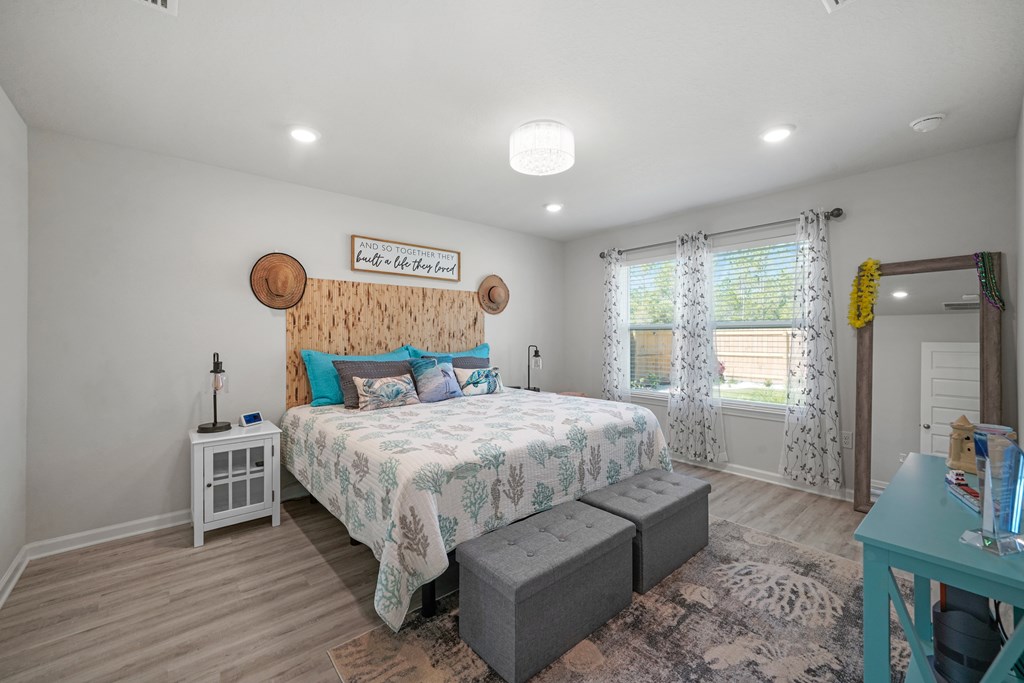 Bedroom with a king-size bed, wood-paneled headboard, gray ottomans, and two curtained windows.