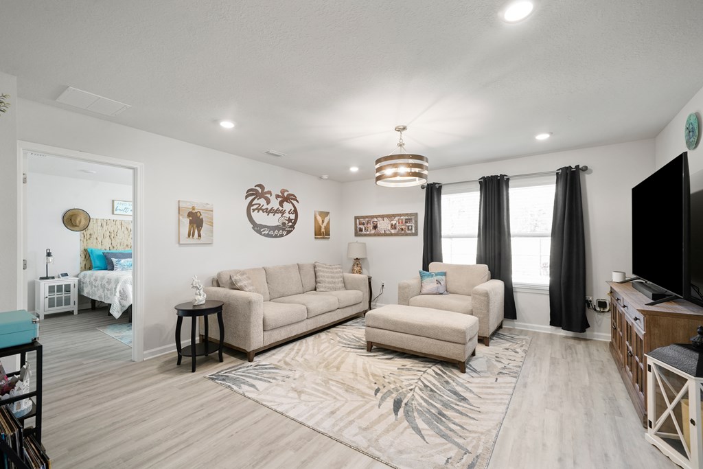 Living room with beige seating, a patterned rug, large curtained windows, and an open bedroom doorway.