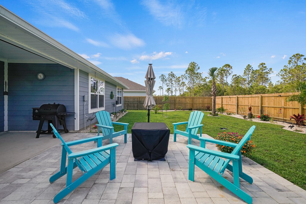 Backyard patio with turquoise Adirondack chairs, covered table, black grill, and fenced lawn.