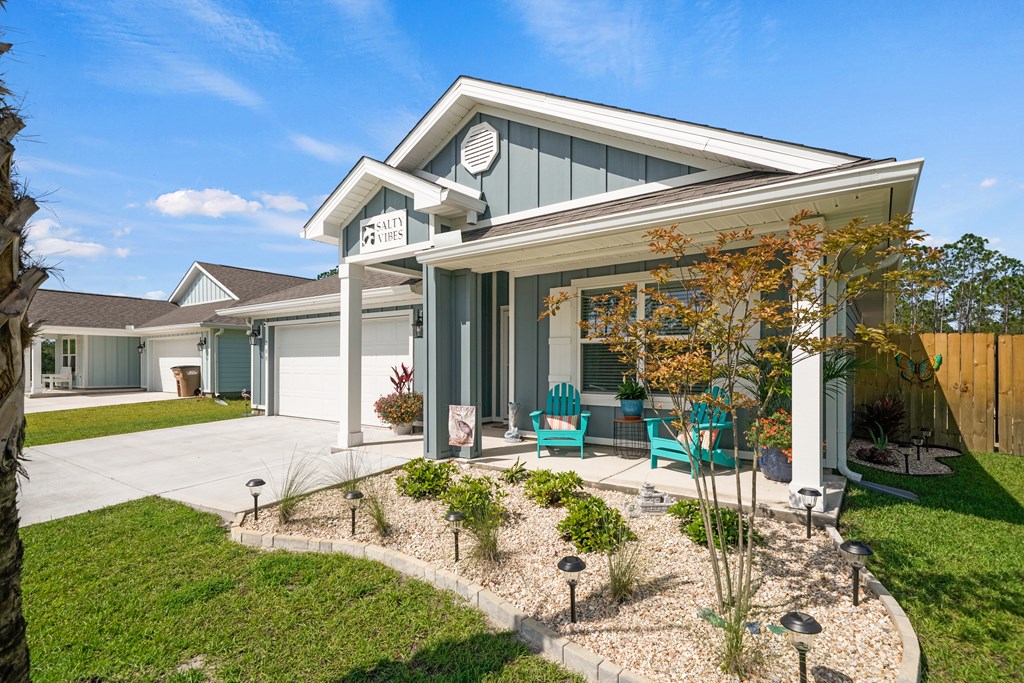 Gray coastal-style home with a covered porch, turquoise chairs, attached garage, and gravel garden.