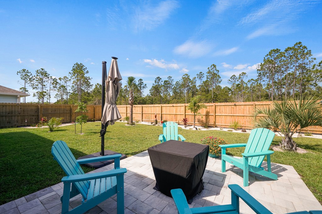 Paver patio with turquoise Adirondack chairs and a covered table beside a fenced lawn.