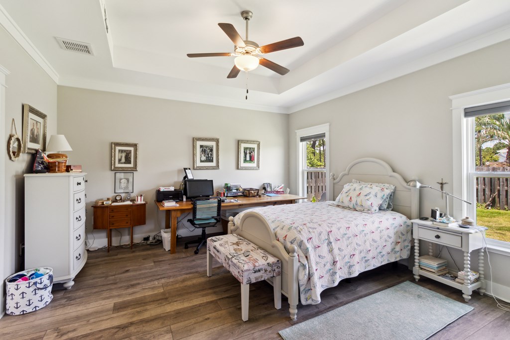 Furnished bedroom with a home office desk, tray ceiling, and two windows overlooking a fenced yard.