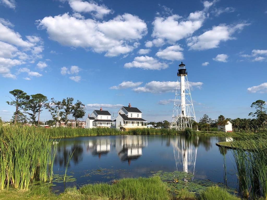 White lighthouse and homes reflected in a pond bordered by reeds.