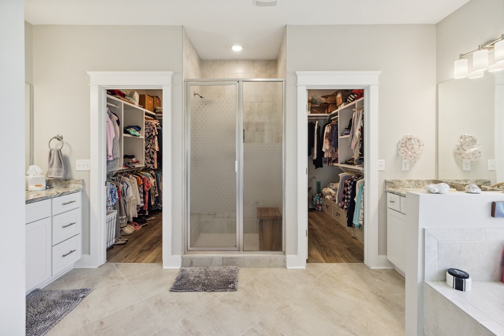 Primary bathroom with a glass-enclosed shower between two walk-in closets and separate vanities.