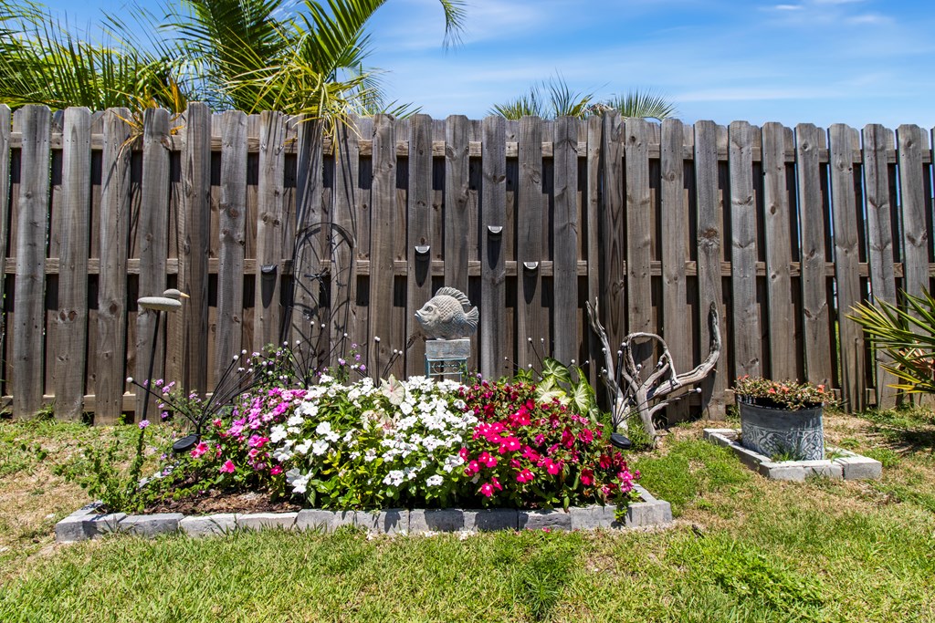 Backyard flower bed with colorful blooms, a fish statue, and a tall wooden privacy fence.
