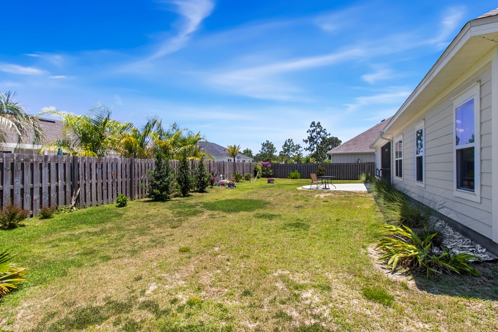 Fenced backyard with lawn, patio seating, and palm landscaping beside the home.