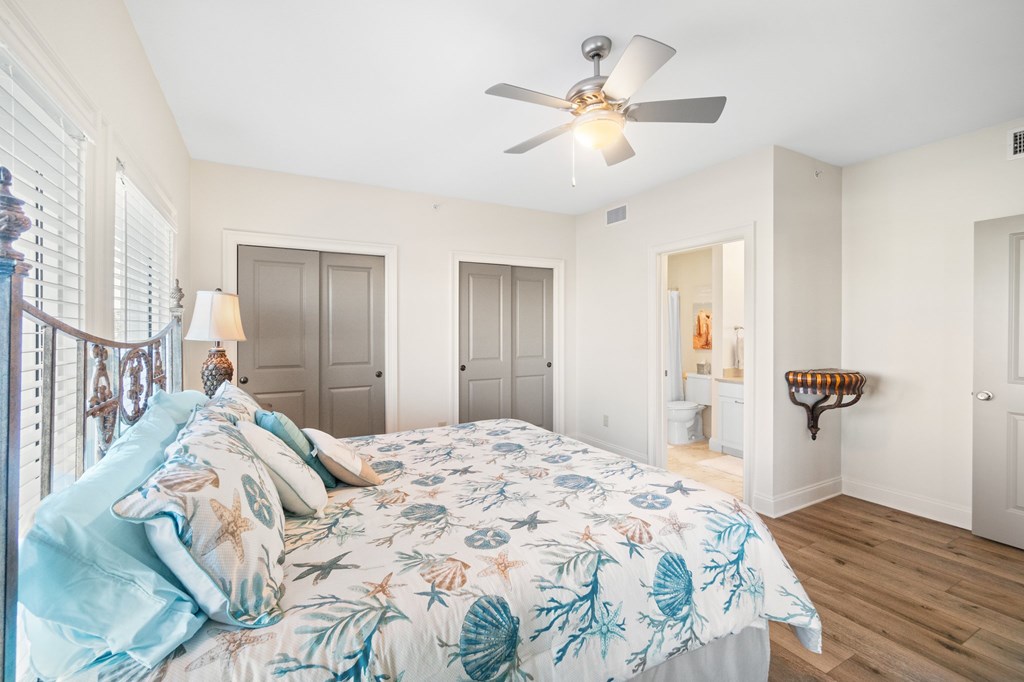 Bedroom with coastal shell-print bedding, plantation shutters, and an adjoining bathroom doorway.