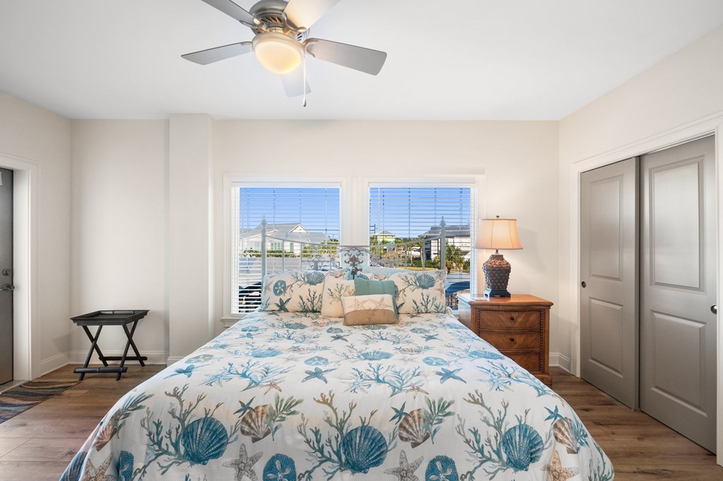 Bedroom with seashell-print bedding, a ceiling fan, and windows overlooking outdoor buildings and greenery.