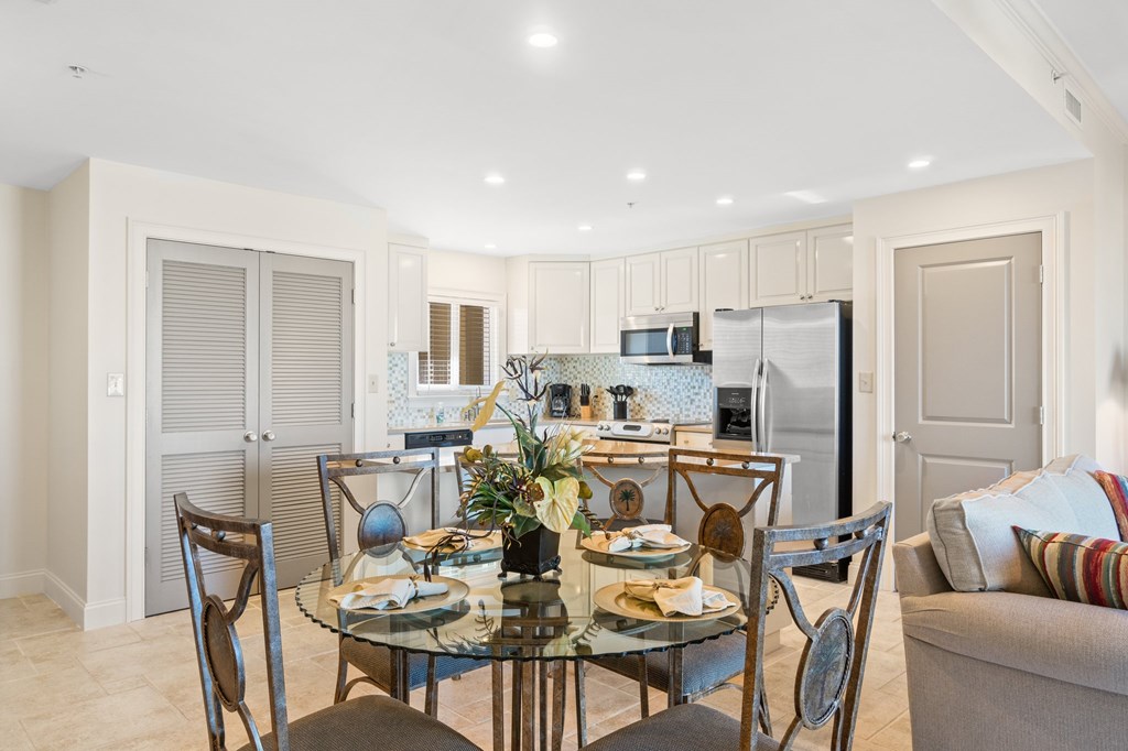 Open kitchen and dining area with a glass-top table, stainless steel appliances, and light tile flooring.