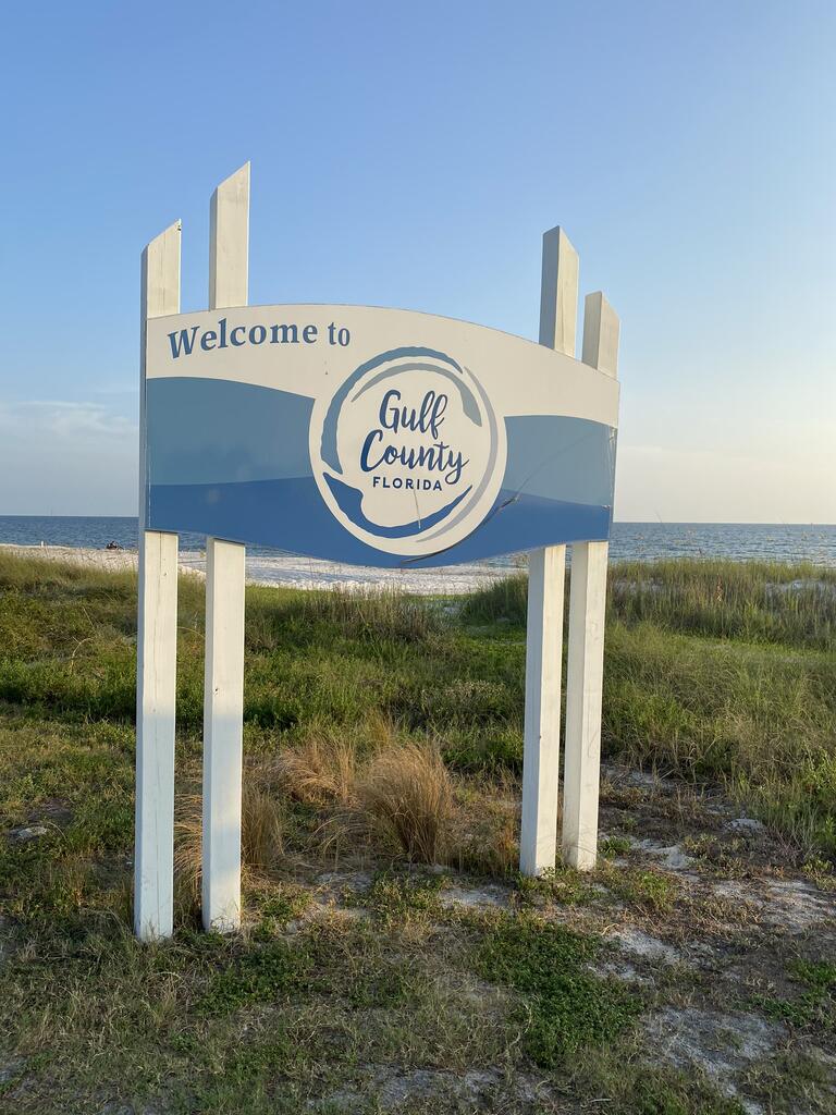 Gulf County welcome sign beside coastal grasses, with the beach and Gulf of Mexico in the background.