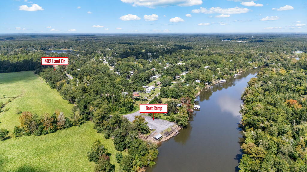 Aerial view of 492 Land Dr beside a river, with a nearby boat ramp and open grassy land.