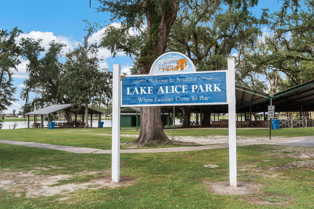 Lake Alice Park welcome sign on a grassy lakeside lawn beneath moss-draped oak trees.