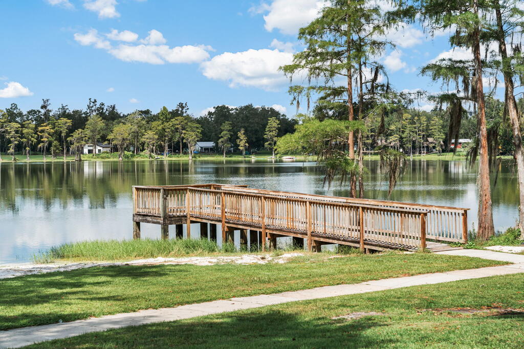 Wooden walkway and deck extending over a calm lake, with trees and homes across the water.
