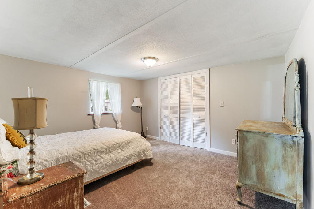 Bedroom with a white quilted bed, curtained window, louvered closet doors, and vintage wooden furniture.