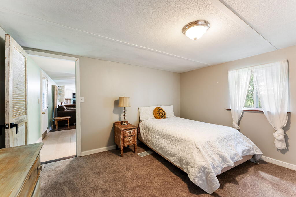 Bedroom with a white-covered bed, wooden nightstand, brown carpet, and curtained window.
