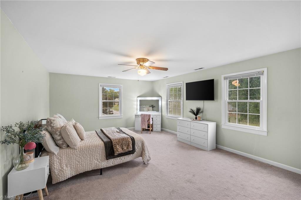 Staged bedroom with a quilted bed, three windows, and a wall-mounted television.