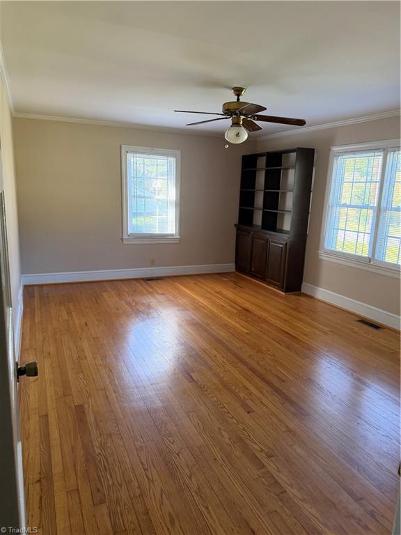 Empty room with hardwood floors, two windows, a ceiling fan, and a built-in bookcase.