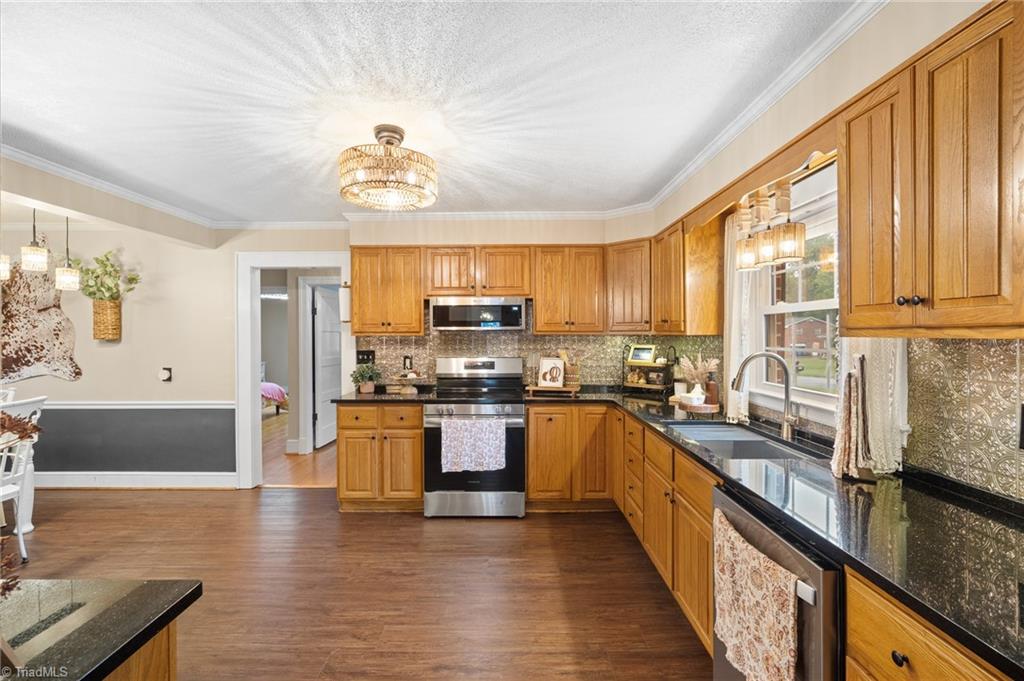 Kitchen with honey-toned wood cabinets, dark countertops, stainless appliances, and a large window above the sink.