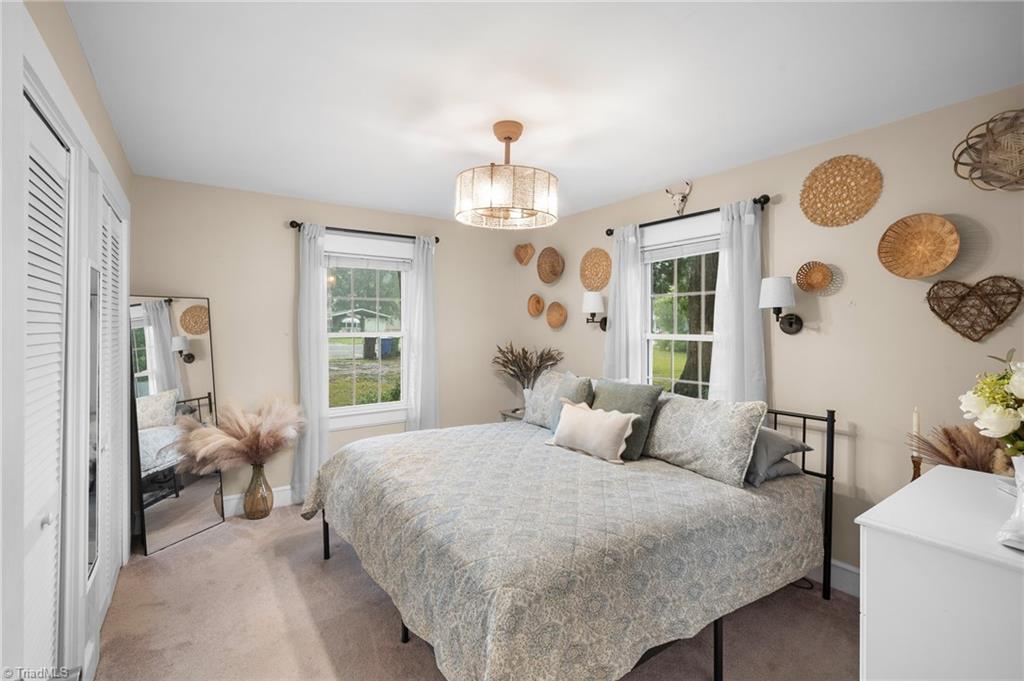 Bedroom with a large pale-covered bed, two curtained windows, woven wall decor, and a chandelier.