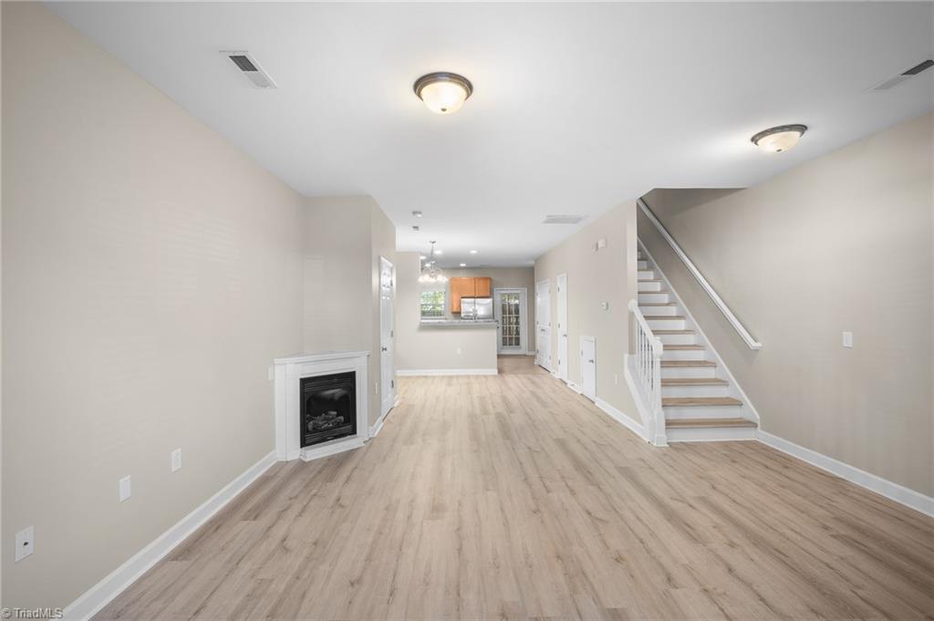 Empty living area with a built-in fireplace, light wood-look floors, and staircase beside the kitchen.