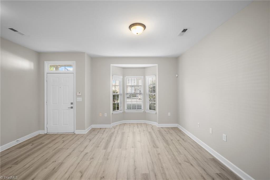 Empty front room with light flooring, an entry door, and a bay window with blinds.