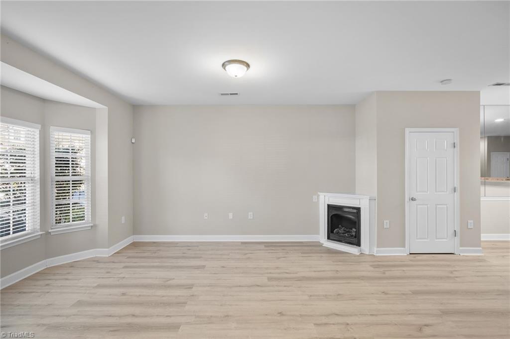 Vacant living room with light wood-look floors, two blind-covered windows, and a corner fireplace.