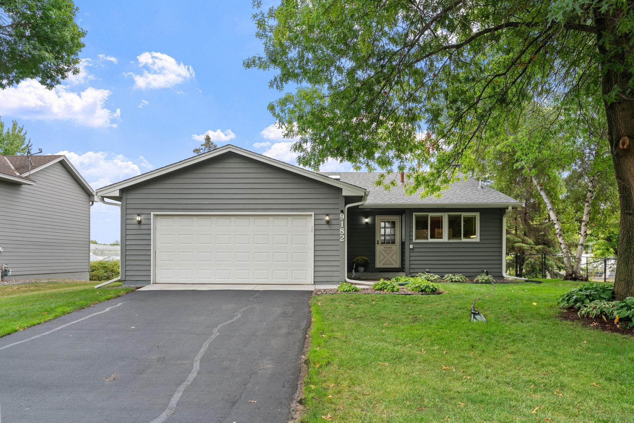 Gray single-story home with a white double garage door, wide driveway, and mature trees.