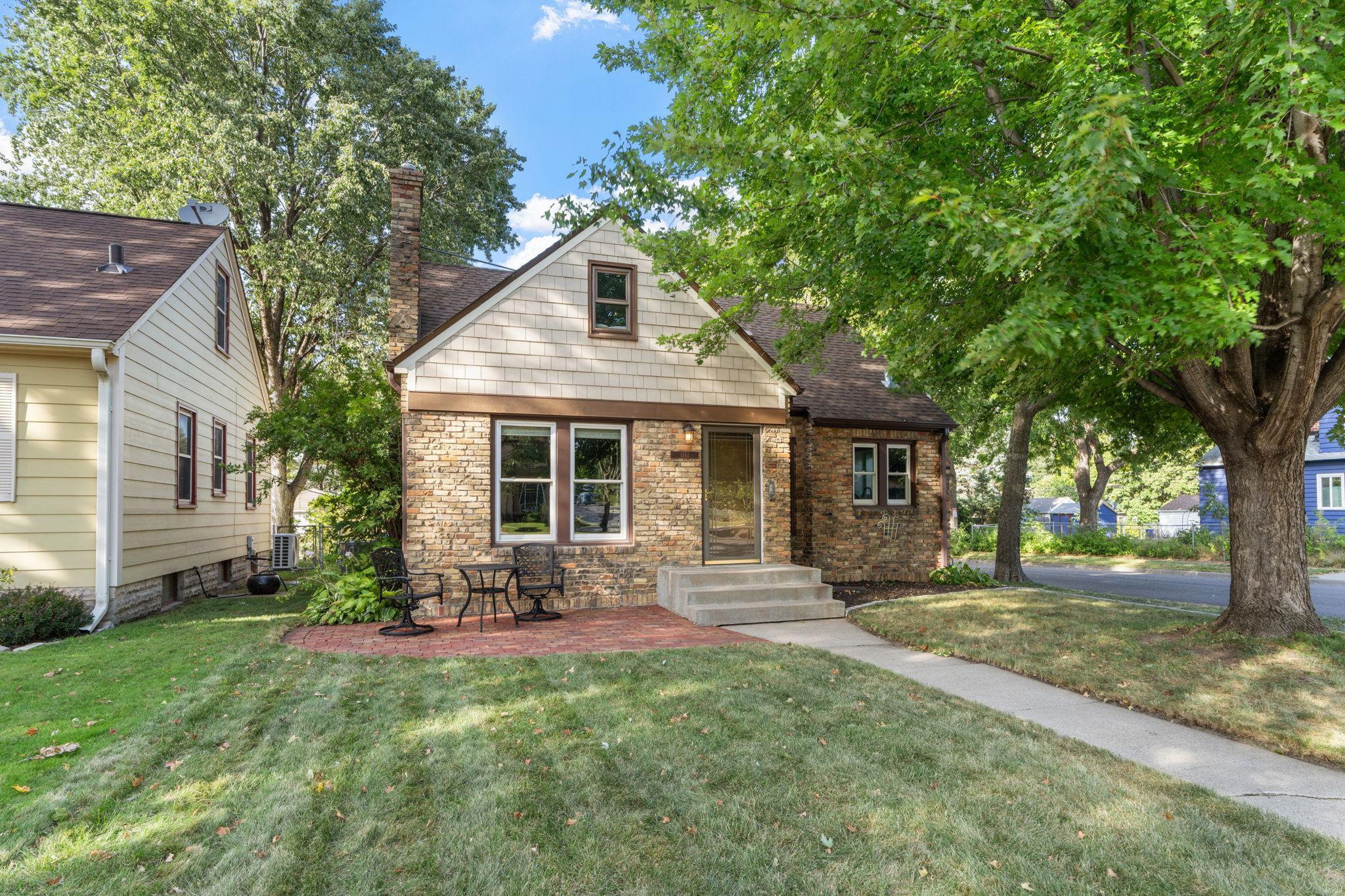 Two-story brick house with a shingled front gable, brick patio, and large shade trees.