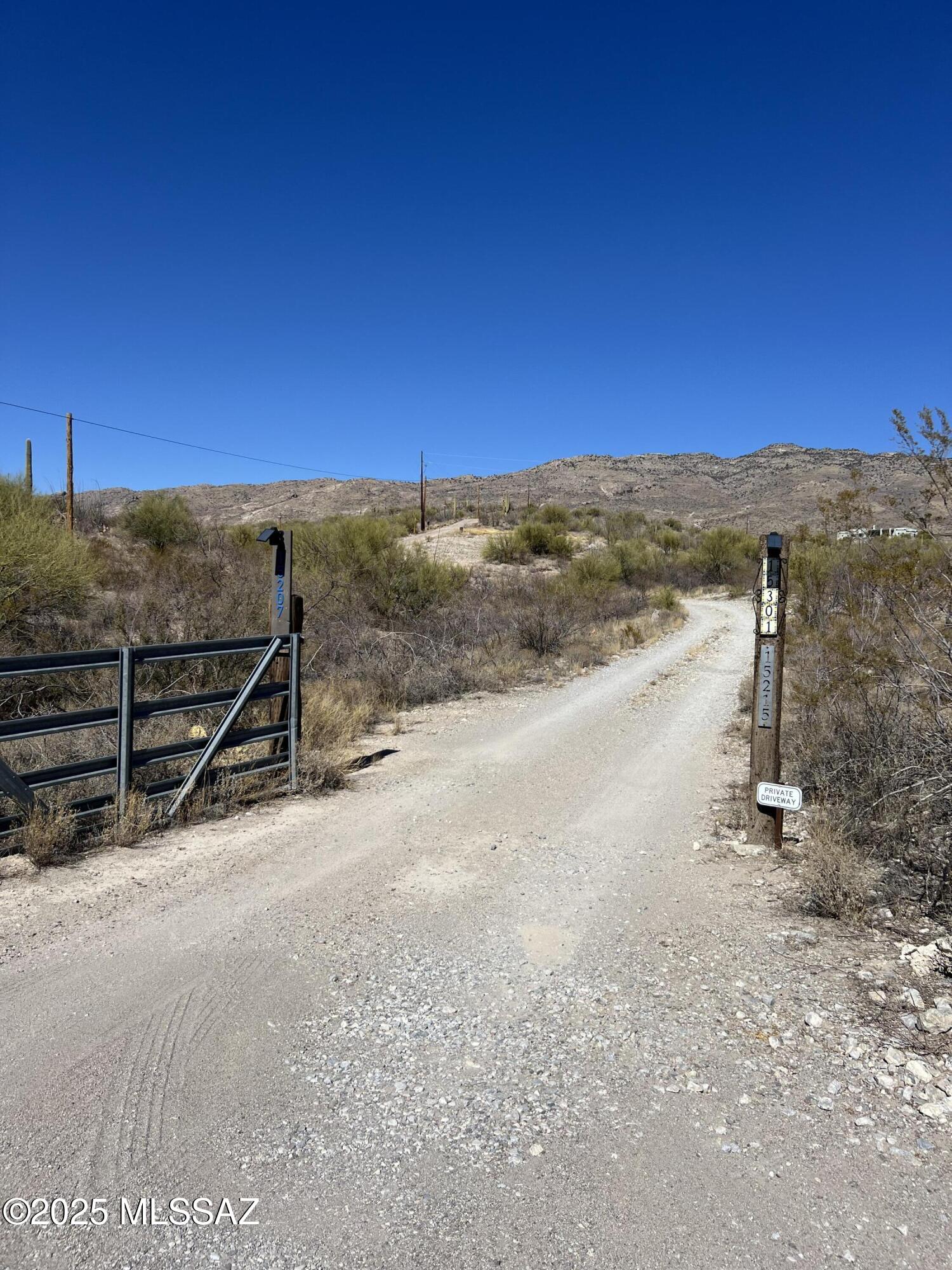 Gravel driveway entrance with an open metal gate, desert brush, and rocky hills beyond.