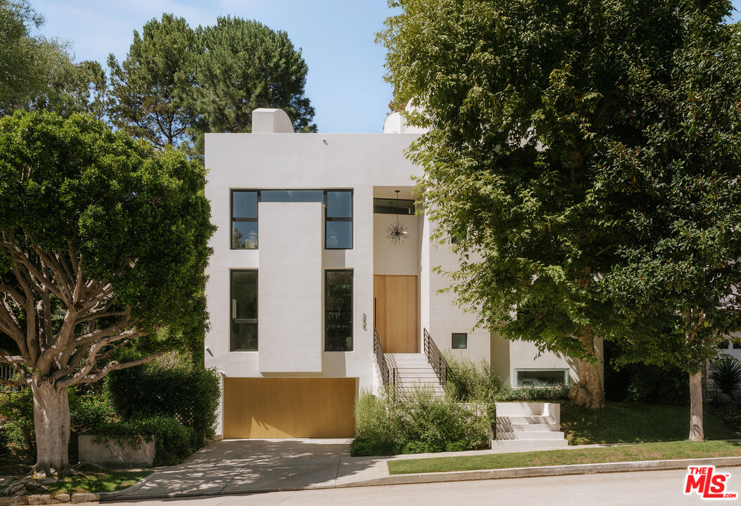 Modern white home with tall windows, a wood-paneled garage, and mature trees framing the entrance.