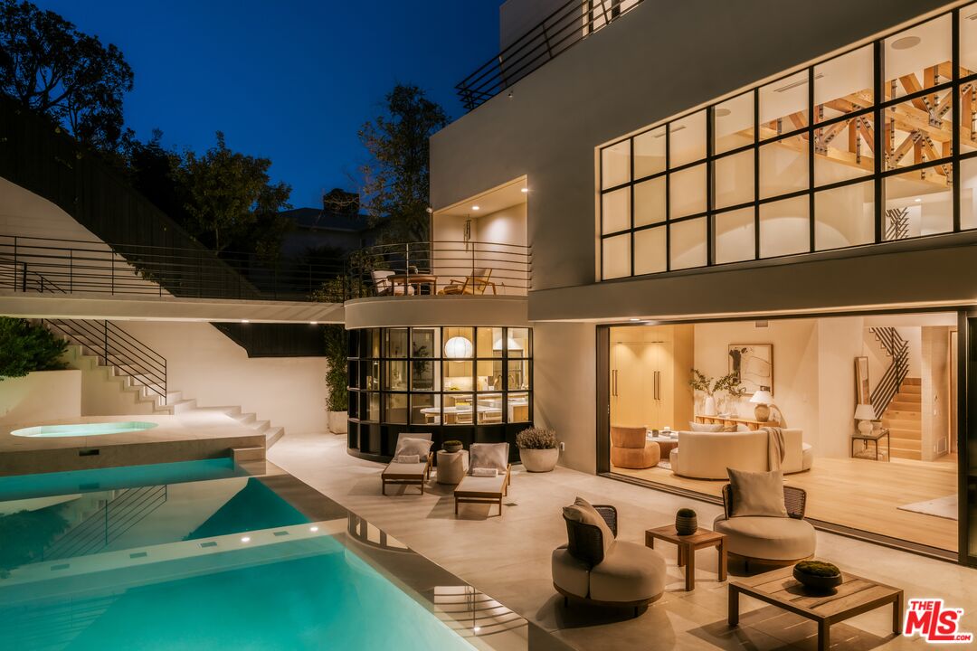 Modern glass-fronted home and illuminated pool at night, with furnished patio seating.