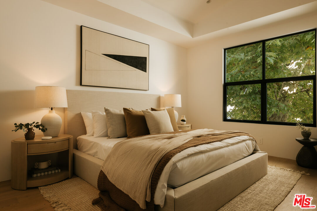 Furnished bedroom with a neutral upholstered bed and a large window overlooking leafy trees.
