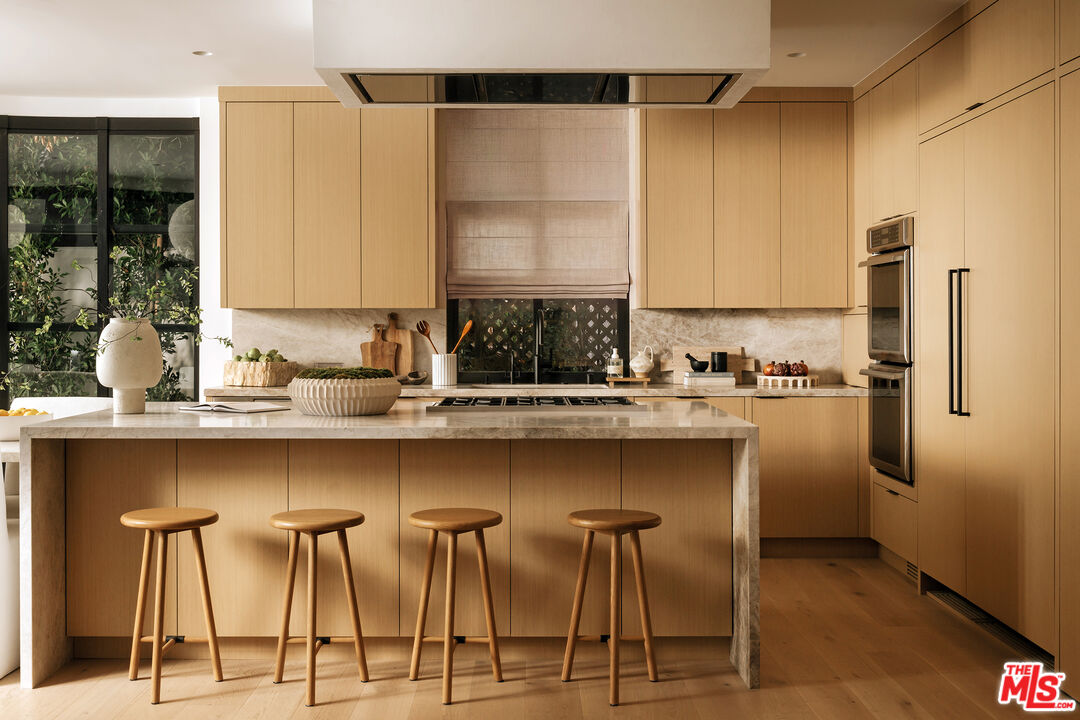 Kitchen with a marble island, four wooden stools, light wood cabinets, and windows overlooking greenery.