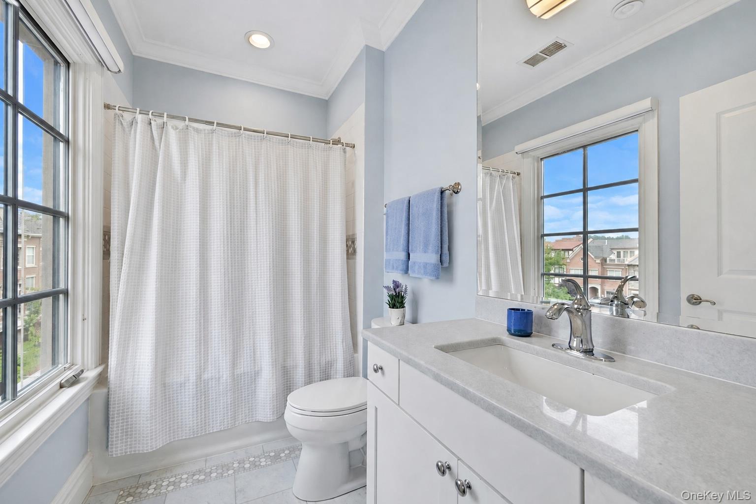 Bathroom with a long white vanity, curtained tub, and tall windows overlooking neighboring buildings.