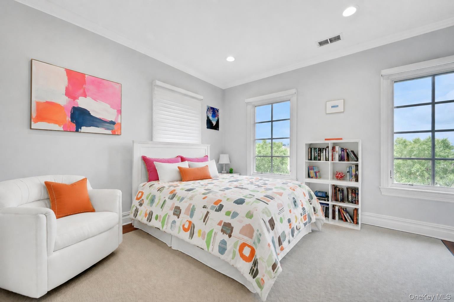 Staged bedroom with a colorful patterned bedspread, white armchair, and tall windows overlooking greenery.