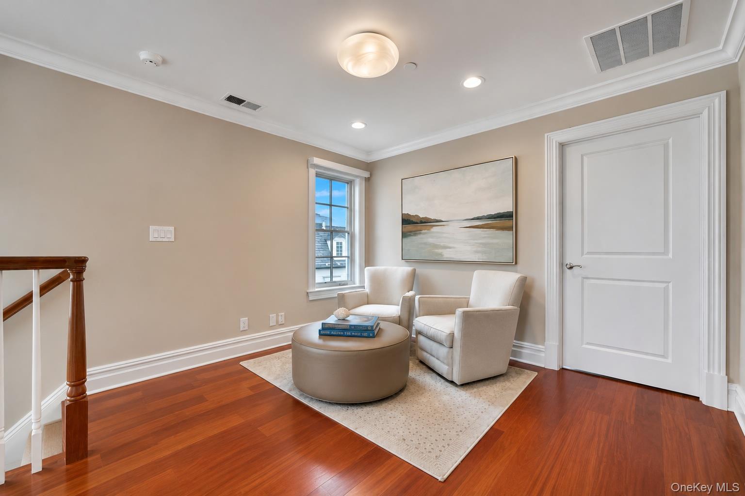 Sitting area with cream armchairs and a round ottoman beside a staircase.