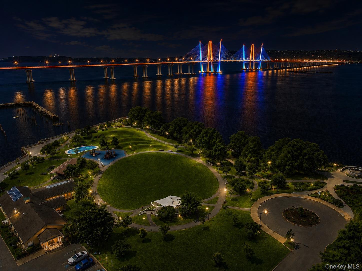An illuminated cable-stayed bridge spans the river above a landscaped park at night.